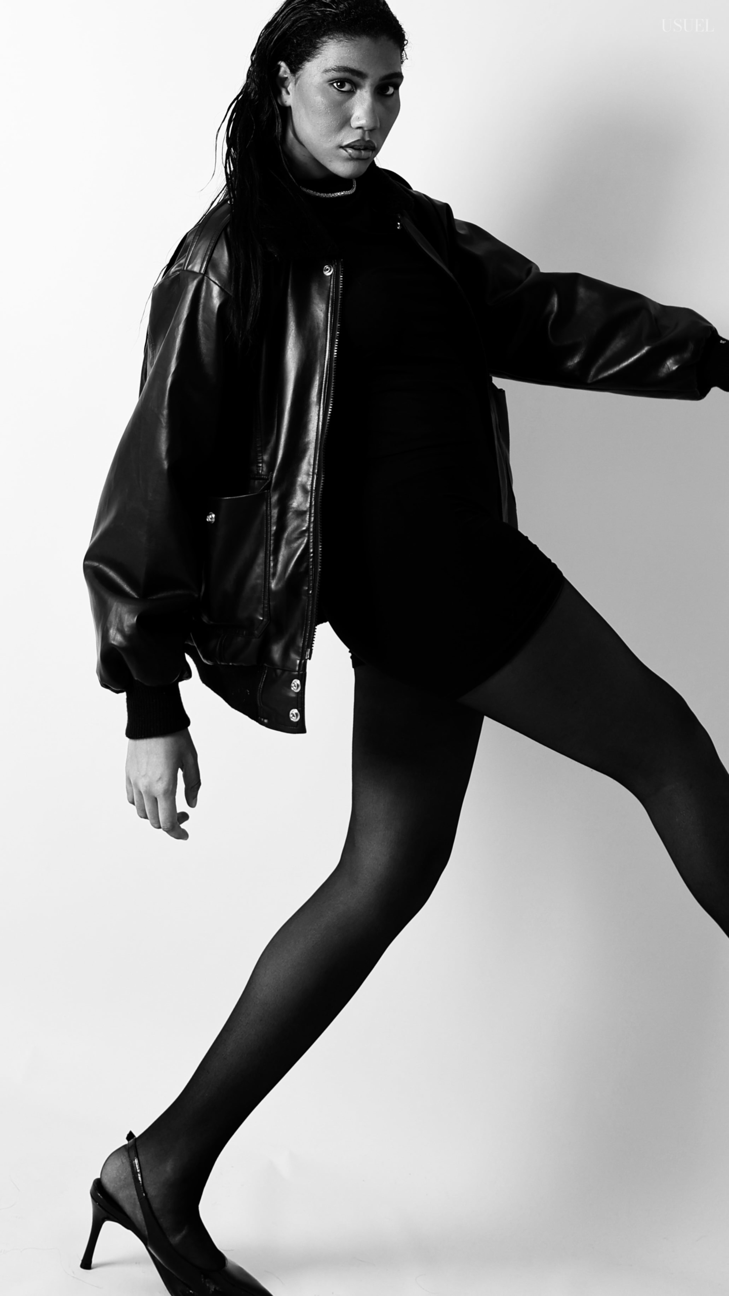 Black and white photo of a woman on a white background, wearing a bold black leather jacket and delicate tights, with an elegant silhouette and confident posture