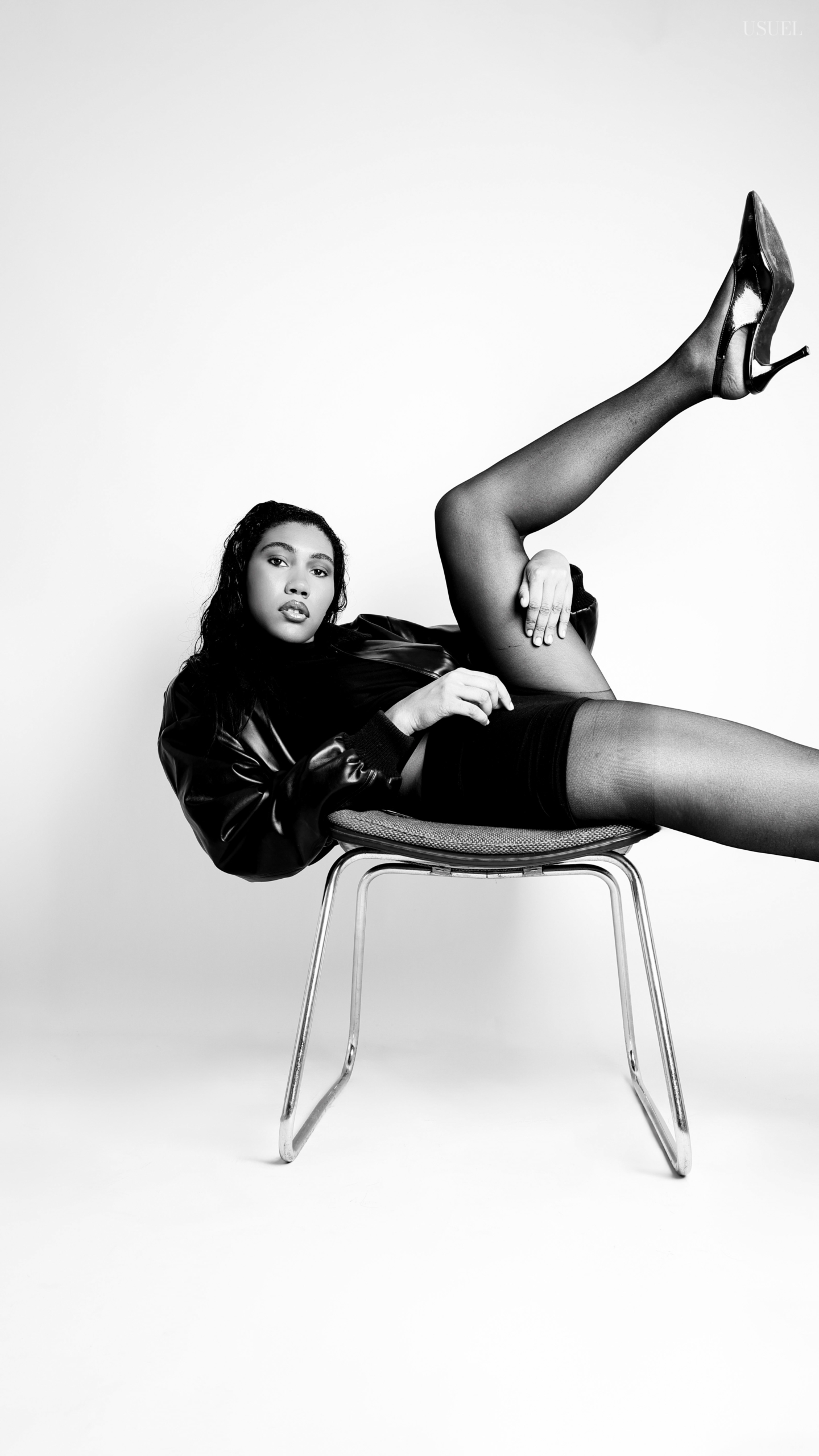 Black and white photo of a woman on a white background, wearing a bold black leather jacket and delicate tights, with an elegant silhouette and confident posture