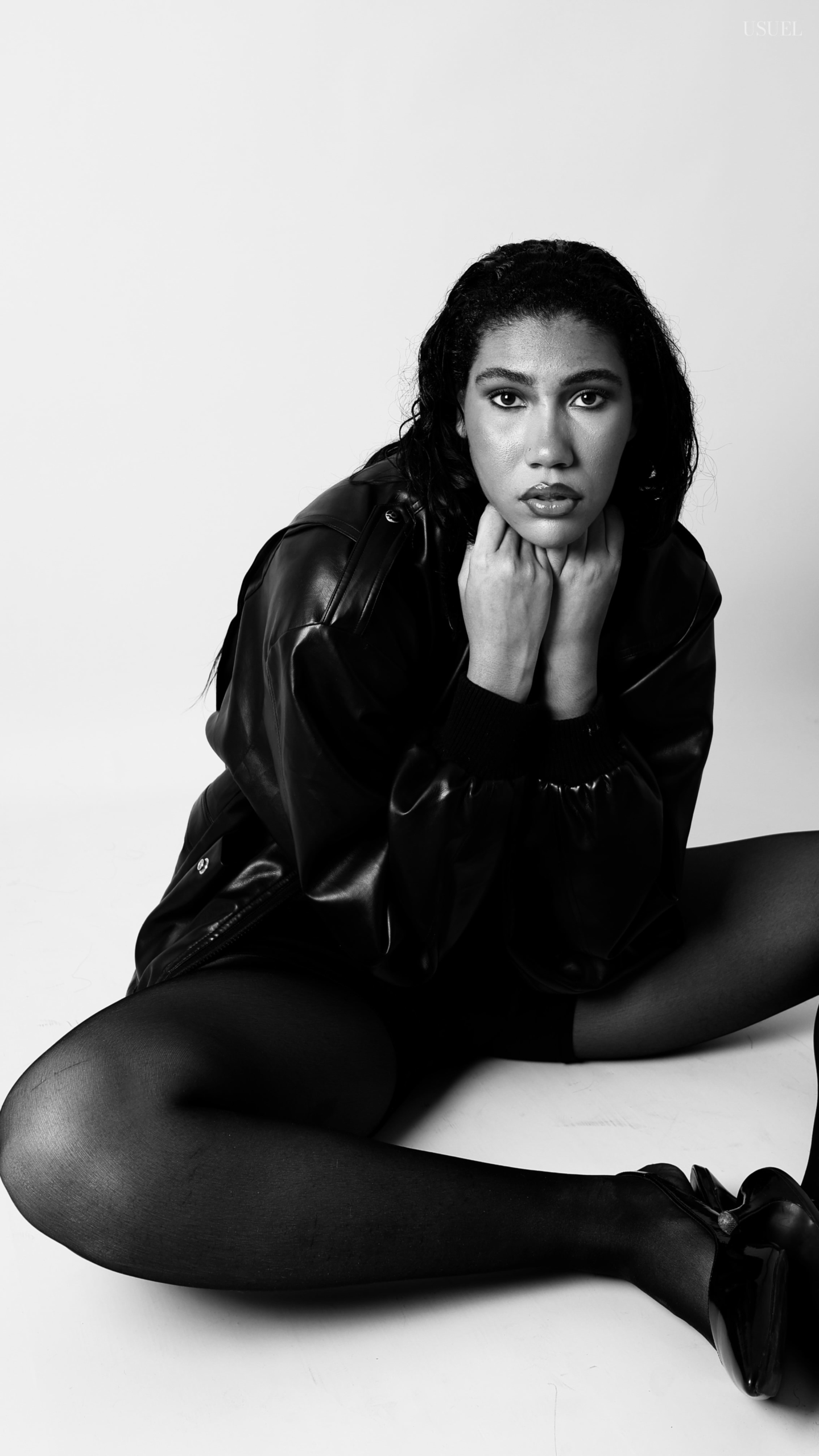 Black and white photo of a woman on a white background, wearing a bold black leather jacket and delicate tights, with an elegant silhouette and confident posture