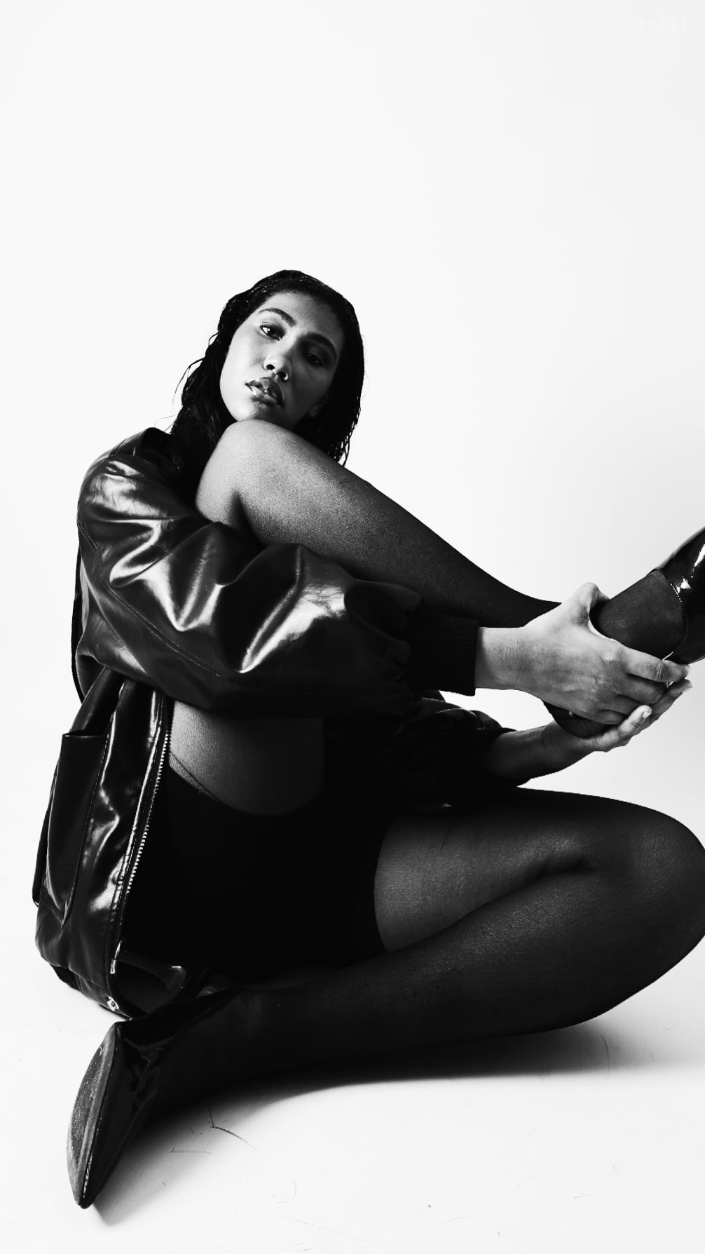 Black and white photo of a woman on a white background, wearing a bold black leather jacket and delicate tights, with an elegant silhouette and confident posture