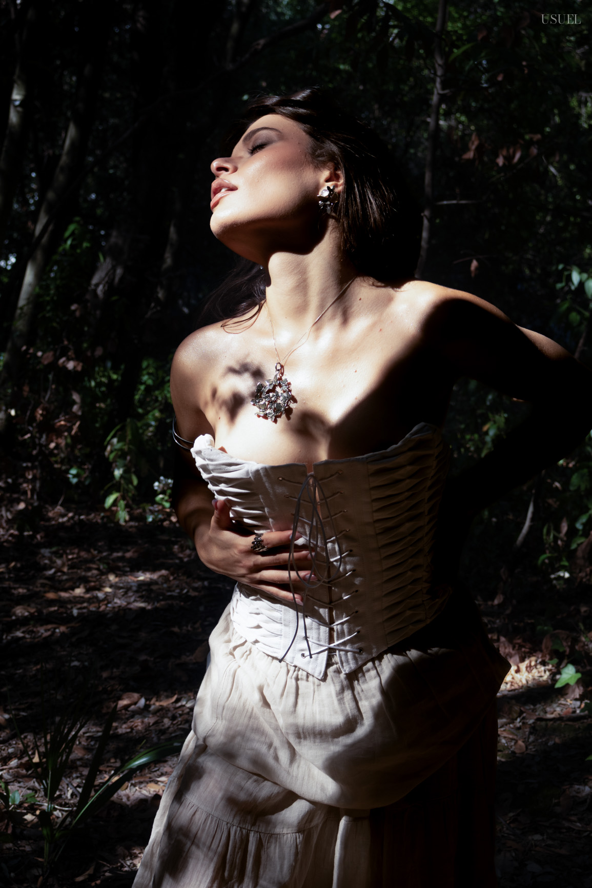 Portrait of a woman wearing a white corset and sculptural pendant, lit by dramatic shadows outdoors