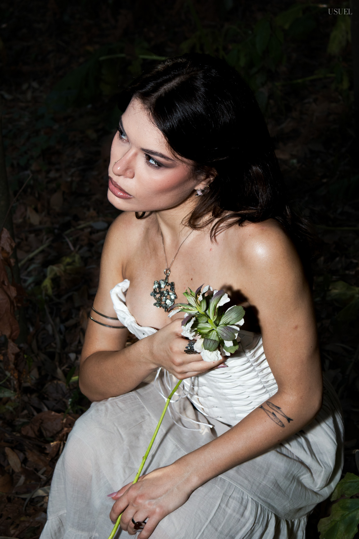 A woman in a white corset kneeling on the forest floor and holding a wild flower