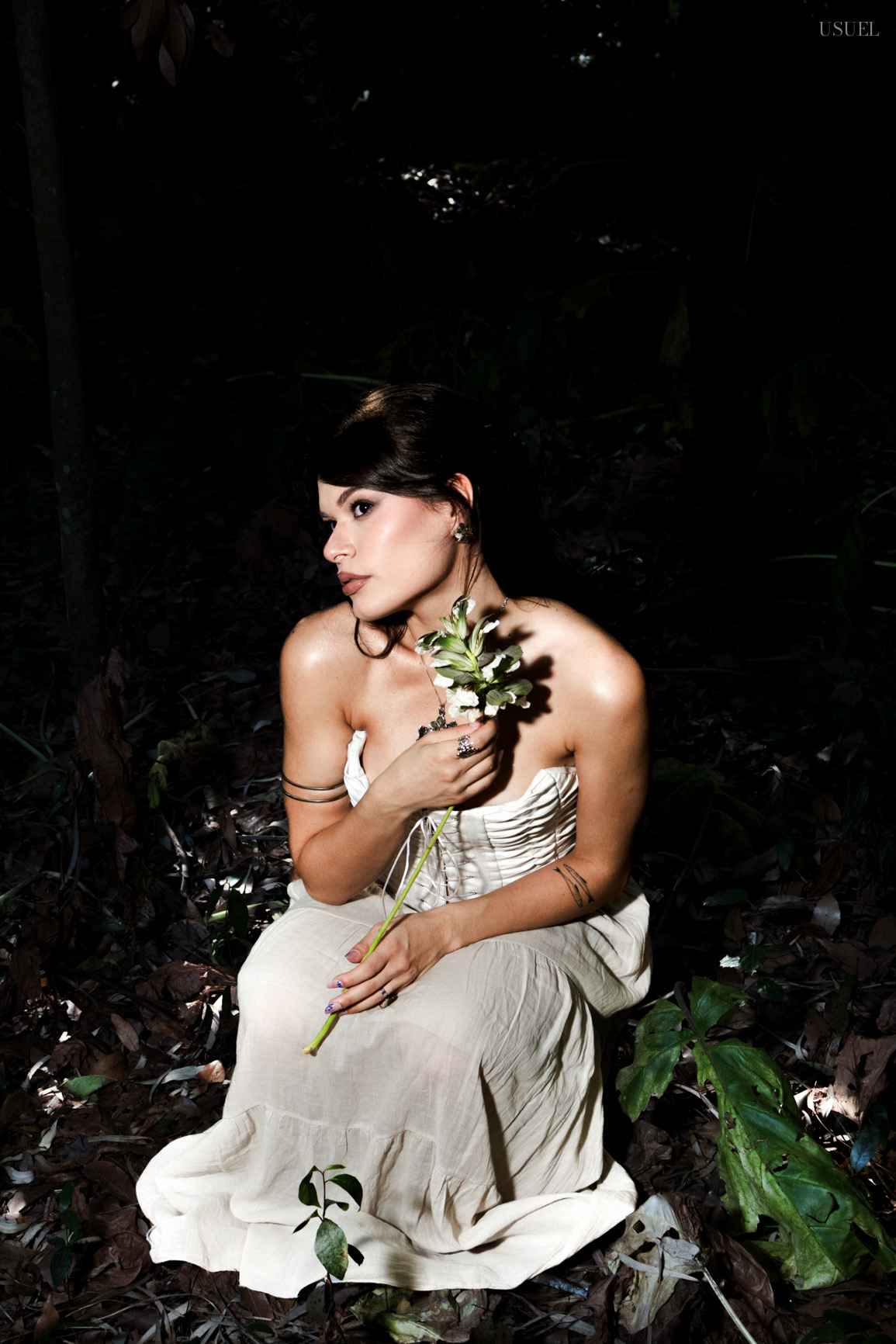 A woman in a white corset kneeling on the forest floor and holding a wild flower