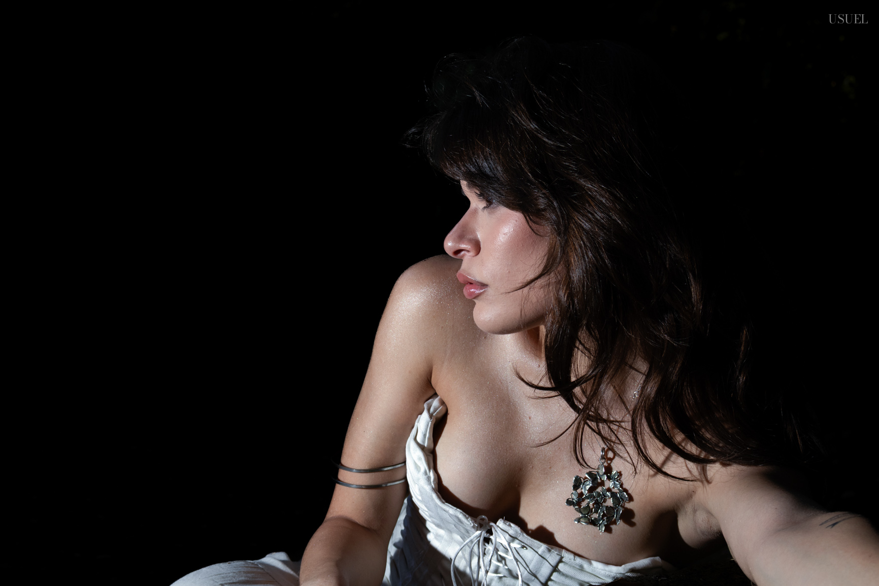 A woman in a white corset lying down on the ground in a dark forest setting, looking away.